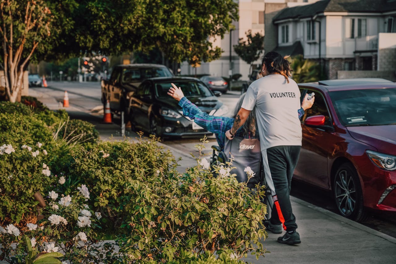 Home Volunteer assisting a person in a wheelchair on a city street surrounded by greenery and parked cars.